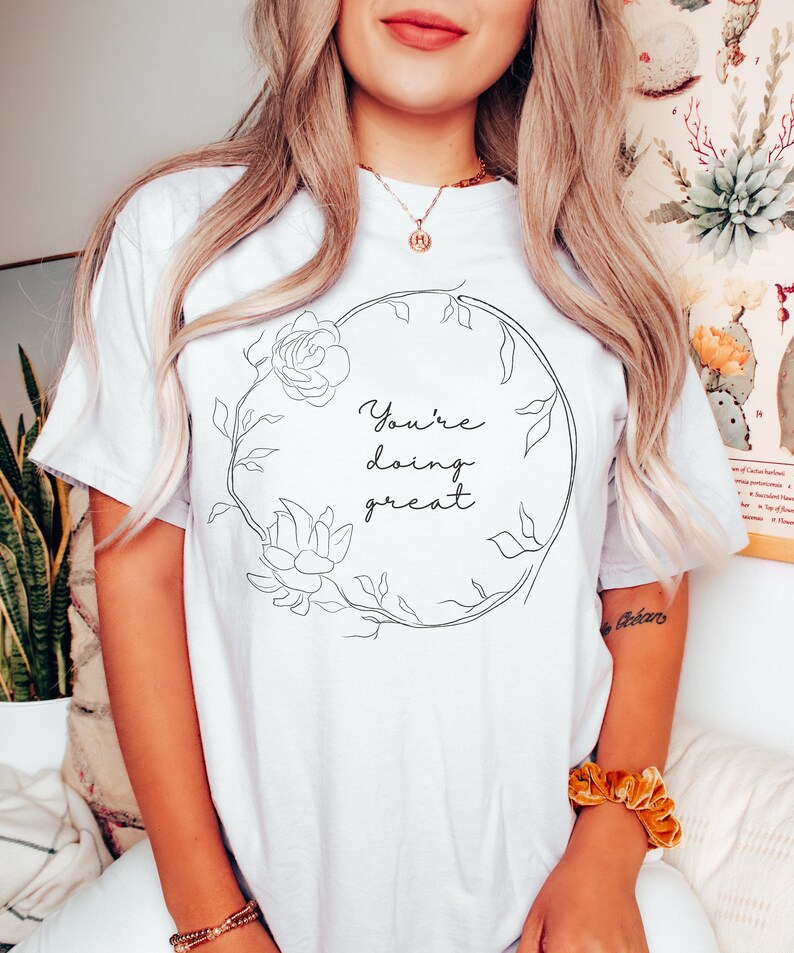 May include: White t-shirt with a floral wreath design and the text "You're doing great" in black script. The wreath features a rose and other floral elements. The shirt is a casual, comfortable style.