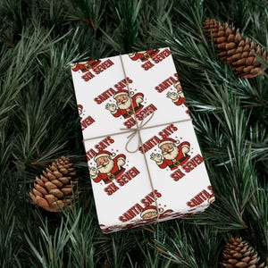 Pode incluir: Presente embrulhado com um tema natalino festivo. O papel de embrulho branco apresenta um padrão repetido de ilustrações do Papai Noel e as frases "SANTA SAYS" e "SIX SEVEN" em vermelho. O presente é amarrado com barbante e colocado sobre agulhas de pinheiro.
