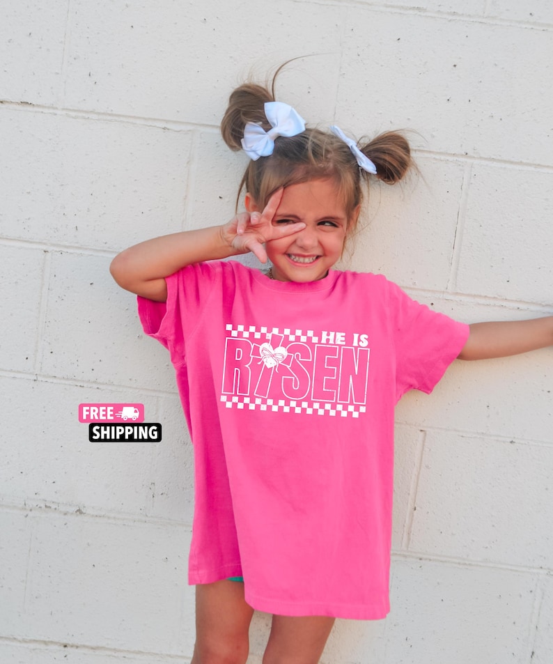 May include: A pink t-shirt with a white graphic that says "He is Risen" with a checkered pattern and a heart with a cross inside.