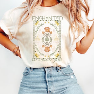 May include: A cream colored t-shirt with a graphic design featuring a floral pattern in shades of green, yellow, and orange. The graphic is surrounded by a border and the text "ENCHANTED to teach you" is printed below the graphic.