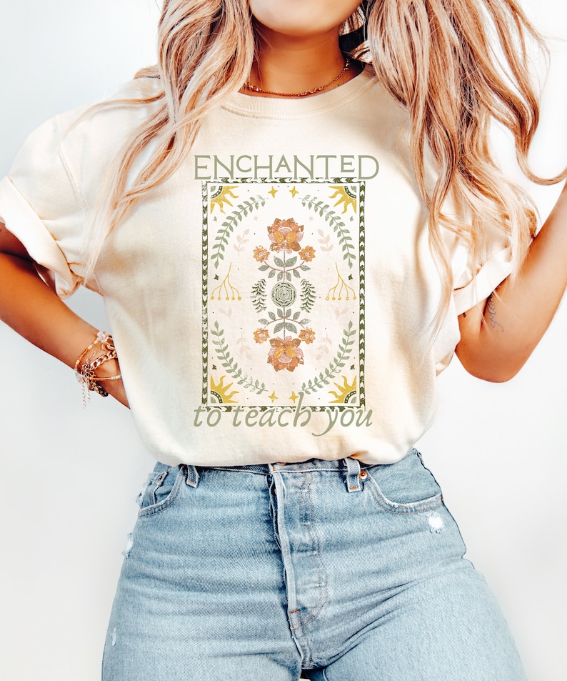 May include: A cream colored t-shirt with a graphic design featuring a floral pattern in shades of green, yellow, and orange. The graphic is surrounded by a border and the text "ENCHANTED to teach you" is printed below the graphic.