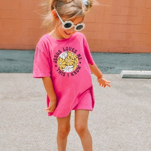 May include: A young girl wearing a pink t-shirt with the text "Jesus Loves Me This I Know" and a circle of yellow flowers. She is wearing white sunglasses and has her hair in pigtails with white bows.