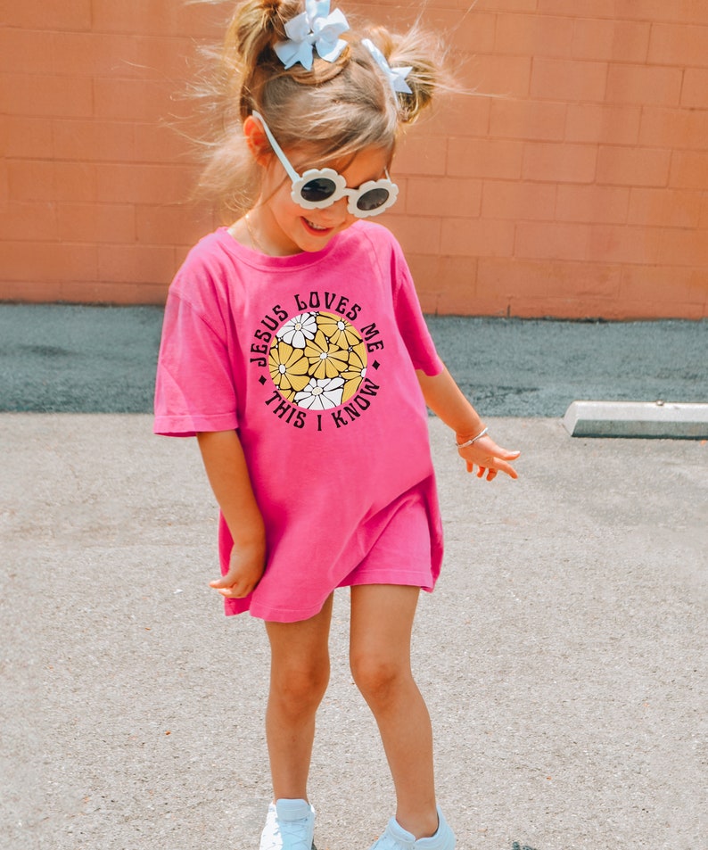 May include: A young girl wearing a pink t-shirt with the text "Jesus Loves Me This I Know" and a circle of yellow flowers. She is wearing white sunglasses and has her hair in pigtails with white bows.