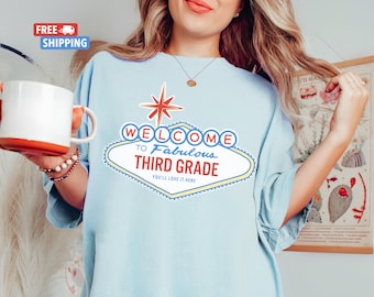 May include: Light blue t-shirt with a graphic of a Las Vegas style sign that says "Welcome to Fabulous Third Grade. You'll Love It Here."