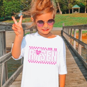 May include: White t-shirt with a pink checkered border and the text "HE IS RISEN" in pink with a pink butterfly graphic.
