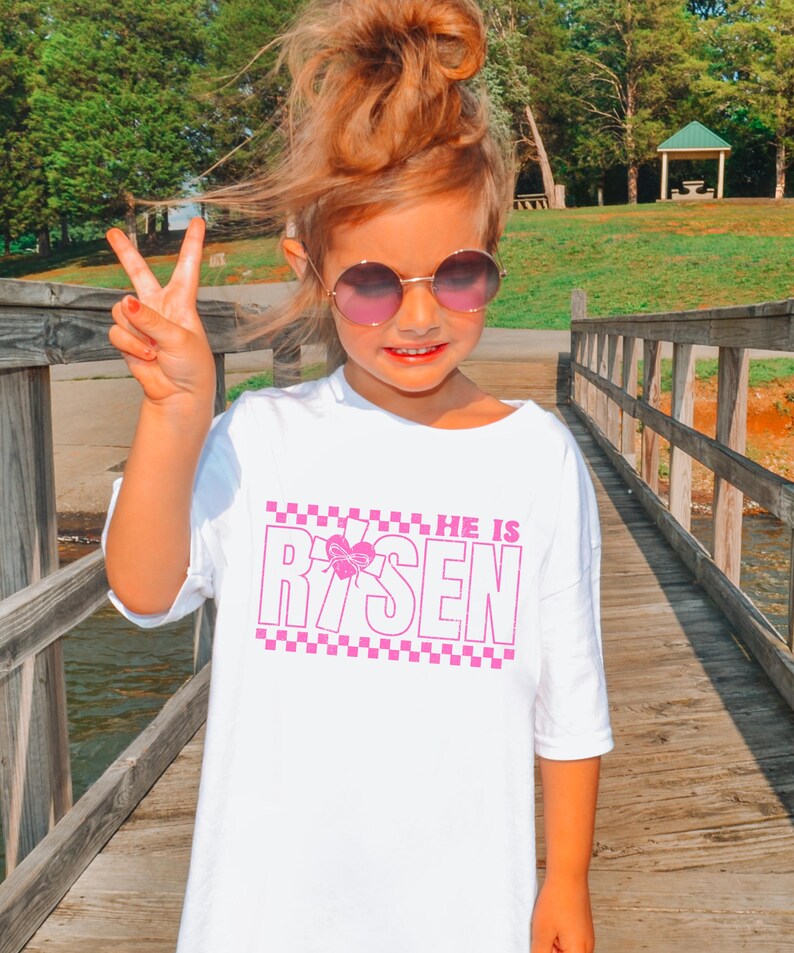 May include: White t-shirt with a pink checkered border and the text "HE IS RISEN" in pink with a pink butterfly graphic.