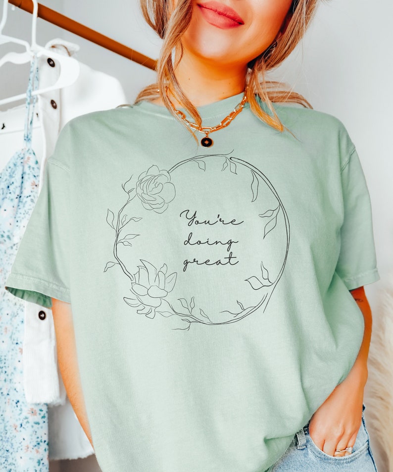 May include: A light green t-shirt with the words "You're doing great" in a floral wreath design. The shirt is a solid color and has short sleeves. The text and floral design are in black.