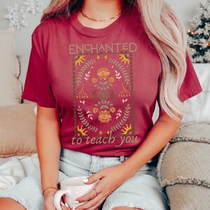 May include: A maroon t-shirt with a floral design and the text "ENCHANTED to teach you".