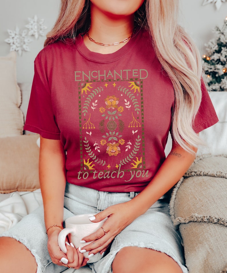 May include: A maroon t-shirt with a floral design and the text "ENCHANTED to teach you".