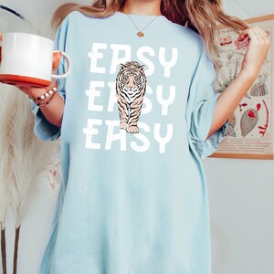 Peut inclure: Un t-shirt surdimensionn&eacute; bleu clair avec un motif blanc d'un tigre marchant avec le mot "EASY" r&eacute;p&eacute;t&eacute; trois fois.