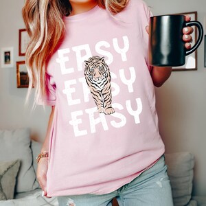 Peut inclure: Un t-shirt rose avec un motif blanc d'un tigre et le mot "EASY" r&eacute;p&eacute;t&eacute; trois fois.