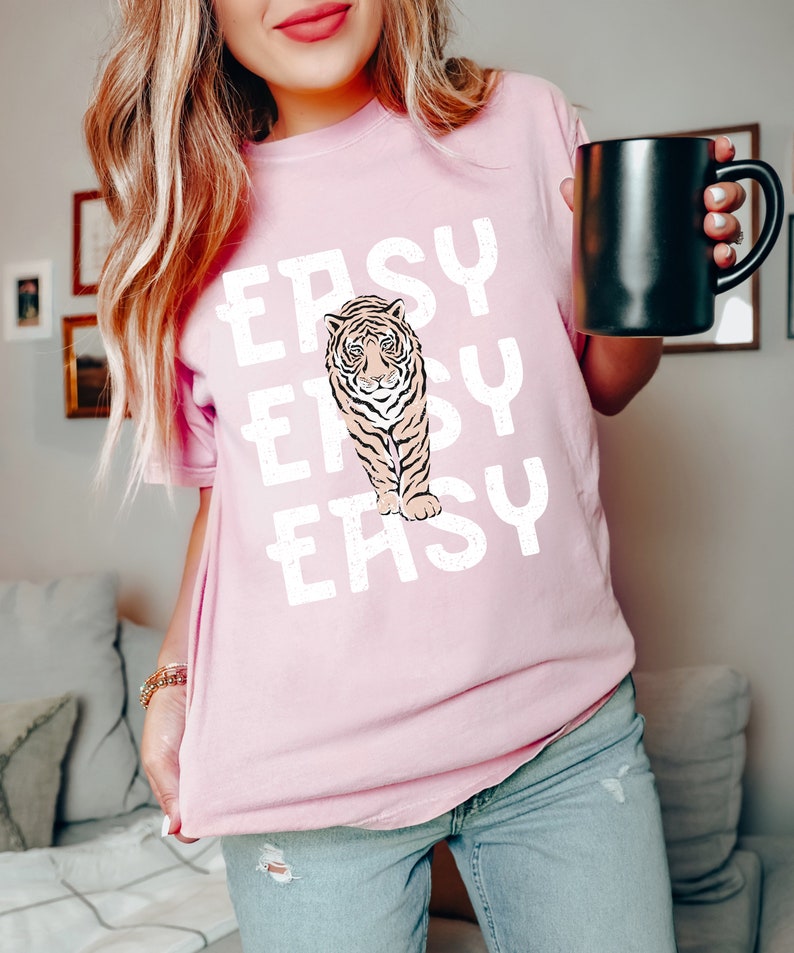 Peut inclure: Un t-shirt rose avec un motif blanc d'un tigre et le mot "EASY" r&eacute;p&eacute;t&eacute; trois fois.