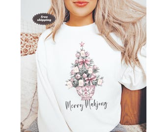 May include: White sweatshirt featuring a Christmas tree design with pink bows, ornaments, and a star. The tree is in a decorative pot, and the words "Merry Mahjong" are written below the tree in a script font. The sweatshirt is a casual style.