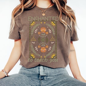 May include: A brown t-shirt with a floral design and the text "ENCHANTED to teach you".