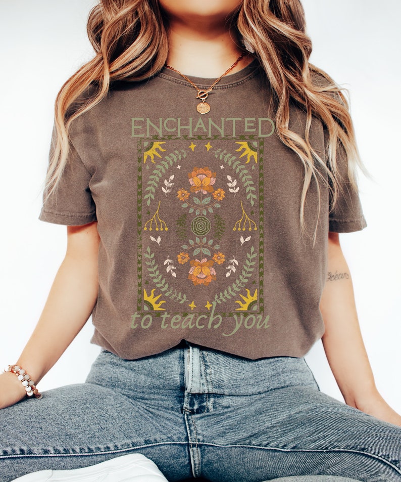 May include: A brown t-shirt with a floral design and the text "ENCHANTED to teach you".