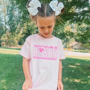 May include: A pink t-shirt with a pink checkered pattern and the words "He is Risen" in pink letters. The shirt has a pink heart with a butterfly design in the middle of the word "Risen".