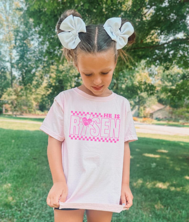 May include: A pink t-shirt with a pink checkered pattern and the words "He is Risen" in pink letters. The shirt has a pink heart with a butterfly design in the middle of the word "Risen".