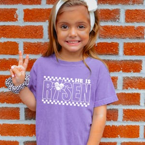 May include: A young girl wearing a purple t-shirt with a white checkered border and the text "HE IS RISEN" with a cross and heart graphic. The girl is smiling and giving a peace sign.