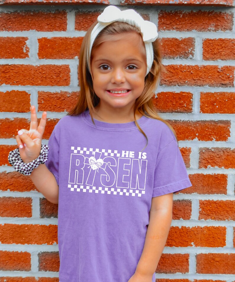 May include: A young girl wearing a purple t-shirt with a white checkered border and the text "HE IS RISEN" with a cross and heart graphic. The girl is smiling and giving a peace sign.