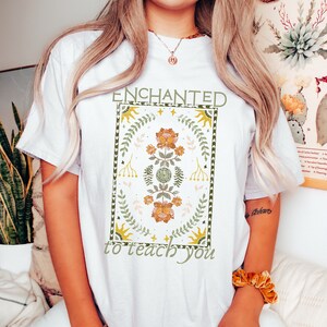 May include: White t-shirt with a floral design and the text "ENCHANTED to teach you".