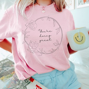 May include: A light pink t-shirt with the text "You're doing great" inside a floral wreath. A white mug with a yellow smiley face is held in the right hand. The person is wearing light blue shorts.