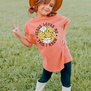 May include: A young girl wearing a coral colored t-shirt with a graphic of a circle with yellow and white flowers and the text "Jesus Loves Me This I Know". She is wearing a brown hat and black leggings with white boots.