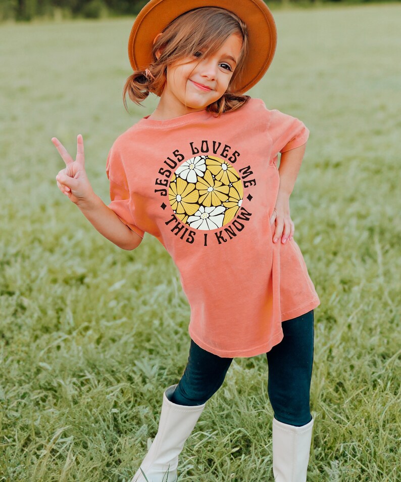 May include: A young girl wearing a coral colored t-shirt with a graphic of a circle with yellow and white flowers and the text "Jesus Loves Me This I Know". She is wearing a brown hat and black leggings with white boots.