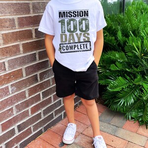 K&ouml;nnte beinhalten: Ein wei&szlig;es T-Shirt mit einer gr&uuml;nen und schwarzen Grafik, auf der "Mission 100 Days Complete" steht. Das Shirt wird von einem Kind getragen, das schwarze Shorts und wei&szlig;e Turnschuhe tr&auml;gt.