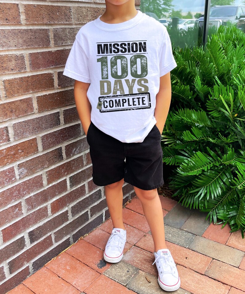 K&ouml;nnte beinhalten: Ein wei&szlig;es T-Shirt mit einer gr&uuml;nen und schwarzen Grafik, auf der "Mission 100 Days Complete" steht. Das Shirt wird von einem Kind getragen, das schwarze Shorts und wei&szlig;e Turnschuhe tr&auml;gt.