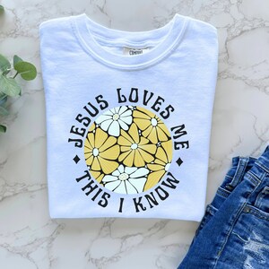 May include: White t-shirt with a circular graphic featuring yellow and white flowers. The graphic has the text "Jesus Loves Me This I Know" in a black circle.