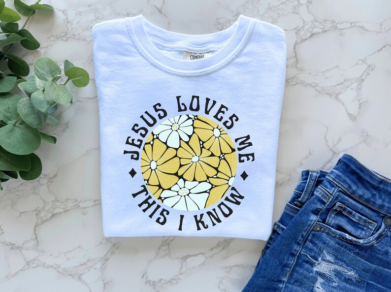 May include: White t-shirt with a circular graphic featuring yellow and white flowers. The graphic has the text "Jesus Loves Me This I Know" in a black circle.