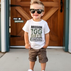 K&ouml;nnte beinhalten: Ein wei&szlig;es T-Shirt mit einer gr&uuml;nen und schwarzen Grafik, auf der "Mission 100 Tage geschafft" steht. Ein kleiner Junge tr&auml;gt das Shirt und dazu braune Shorts und wei&szlig;e Sneaker. Er tr&auml;gt eine Sonnenbrille.
