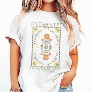 May include: White t-shirt with a graphic design featuring a floral pattern in shades of green, yellow, and pink. The graphic is surrounded by a border and the text "ENCHANTED to teach you" is printed below the graphic.