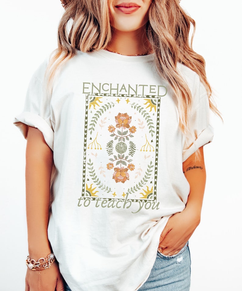 May include: White t-shirt with a graphic design featuring a floral pattern in shades of green, yellow, and pink. The graphic is surrounded by a border and the text "ENCHANTED to teach you" is printed below the graphic.