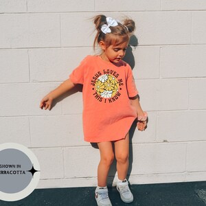 May include: A young girl wearing a terracotta orange t-shirt with a white daisy graphic and the text "Jesus Loves Me This I Know".