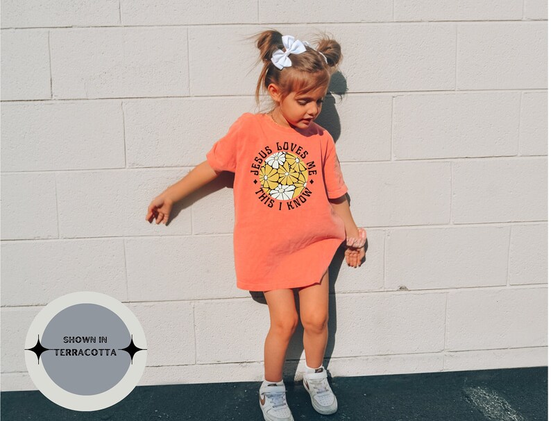 May include: A young girl wearing a terracotta orange t-shirt with a white daisy graphic and the text "Jesus Loves Me This I Know".