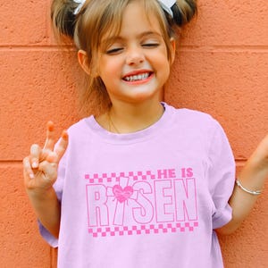 May include: A pink t-shirt with a pink graphic that says "HE IS RISEN" with a pink heart and cross design. The shirt has a checkered pattern at the bottom.