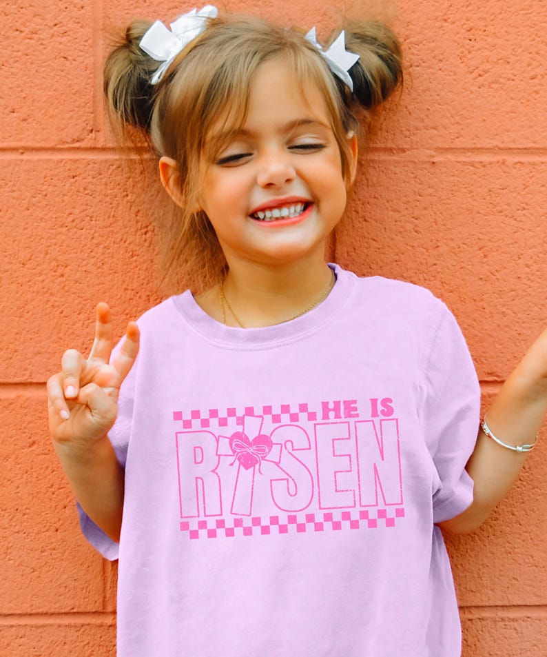 May include: A pink t-shirt with a pink graphic that says "HE IS RISEN" with a pink heart and cross design. The shirt has a checkered pattern at the bottom.