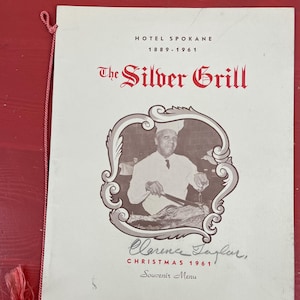 May include: A black and white photo of a chef in a white uniform and chef's hat carving a roast. The photo is framed with a decorative border and the text "The Silver Grill" is printed in red above the photo. The text "Hotel Spokane 1889-1961" is printed above the title. The text "Christmas 1961 Souvenir Menu" is printed below the photo. The chef's signature "Clarence Taylor" is printed below the text.