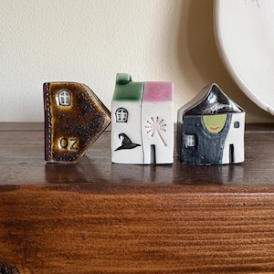 May include: Three ceramic house figurines. The first is brown with "OZ" text. The second is white and pink with a witch hat and a starburst. The third is black and white with a smiling face. They are displayed on a wooden shelf.