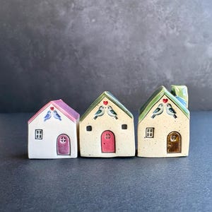 Może przedstawiać: Trzy ceramiczne domki z ręcznie malowanymi detalami. Każdy domek ma inny kolor dachu, drzwi i okna. Domki zdobione są motywami ptaków i serc. Domki ustawione są w rzędzie na ciemnym tle.