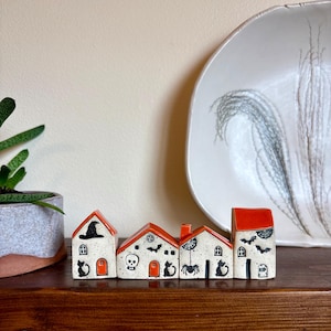 May include: Four small ceramic house figurines with speckled white surfaces and bright orange roofs. Each house features black hand-painted details including cats, a witch hat, a skull, bats, a spider with a web, and a tombstone with the text RIP.