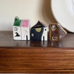 May include: Three ceramic houses with whimsical designs. One house has a green and pink roof with a witch's hat and a starburst. Another is black and white with a smiling face. The last is brown with the text "OZ".