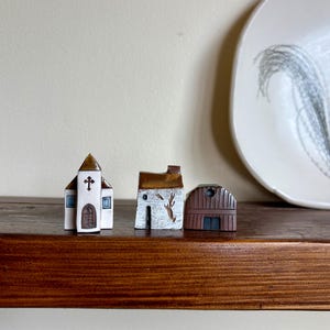 Puede incluir: Tres edificios de cerámica en miniatura: una iglesia, una casa y un granero. La iglesia tiene una cruz en la parte superior y una puerta marrón. La casa tiene un diseño de árbol en el lateral. El granero es marrón con una puerta azul oscuro. Se exhiben en una estantería de madera.