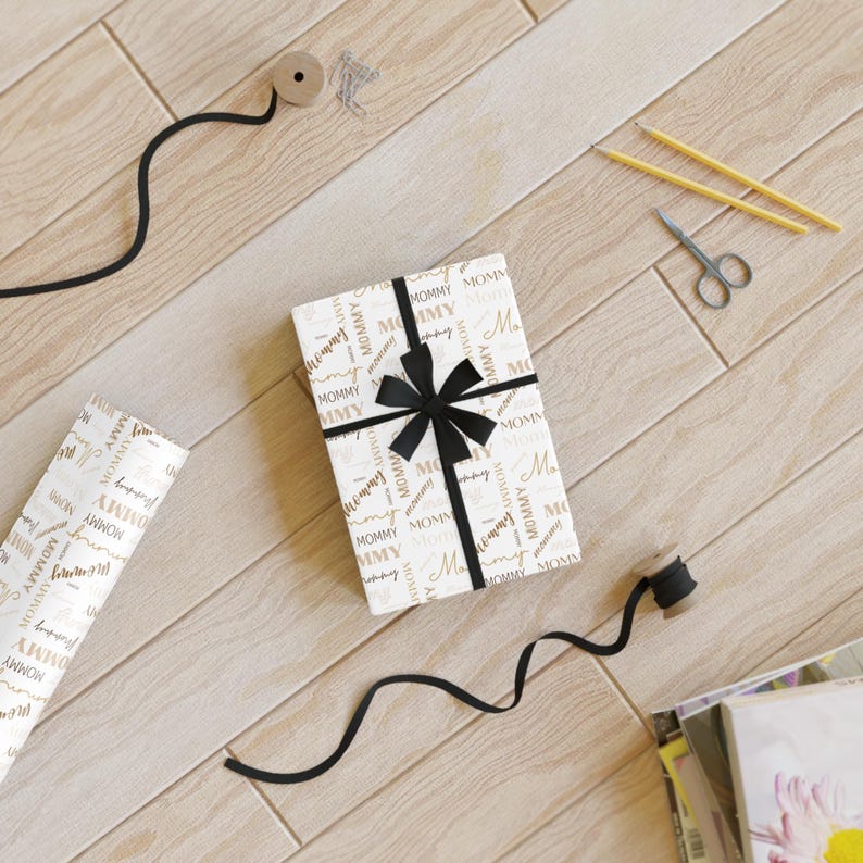 Puede incluir: Un papel de regalo blanco con un patr&oacute;n dorado y negro. El patr&oacute;n presenta la palabra "Mommy" repetida a lo largo. Una cinta negra est&aacute; atada alrededor del papel de regalo con un lazo.