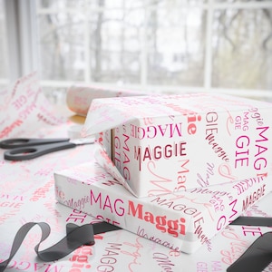 Puede incluir: Una caja de regalo blanca envuelta en papel estampado rosa y blanco con el nombre "Maggie" repetido en rosa. La caja est&aacute; encima de otra caja de regalo envuelta en el mismo papel.