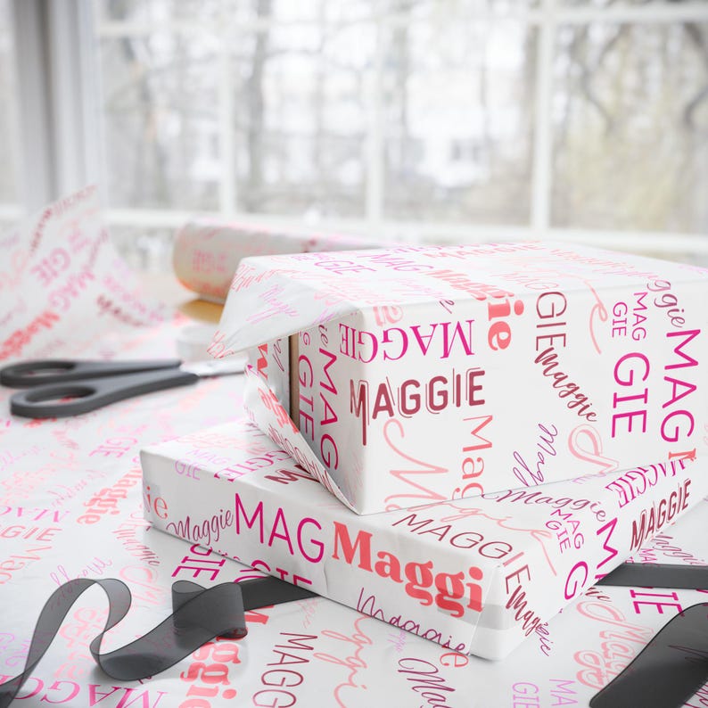 Puede incluir: Una caja de regalo blanca envuelta en papel estampado rosa y blanco con el nombre "Maggie" repetido en rosa. La caja est&aacute; encima de otra caja de regalo envuelta en el mismo papel.