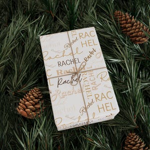 Puede incluir: Papel de regalo blanco con el nombre "Rachel" impreso en letras doradas de escritura y en letras may&uacute;sculas. El papel est&aacute; atado con un lazo de cuerda marr&oacute;n.