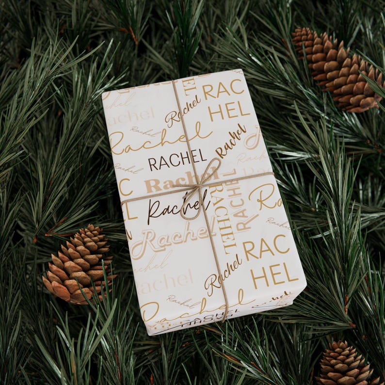 Puede incluir: Papel de regalo blanco con el nombre "Rachel" impreso en letras doradas de escritura y en letras may&uacute;sculas. El papel est&aacute; atado con un lazo de cuerda marr&oacute;n.