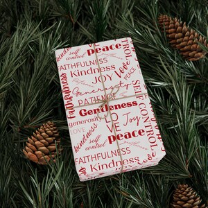 Puede incluir: Papel de regalo blanco con texto rojo que incluye palabras como "paz", "amor", "alegr&iacute;a", "amabilidad", "fidelidad" y "generosidad". El regalo est&aacute; envuelto con cuerda y se puede ver un peque&ntilde;o cono de pino marr&oacute;n en el fondo.